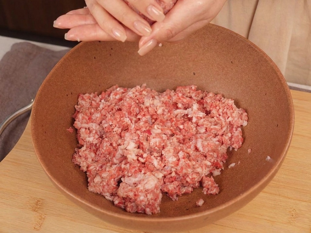 Le mani di una persona che modellano una palla tonda di carne macinata cruda sopra una ciotola di ceramica.