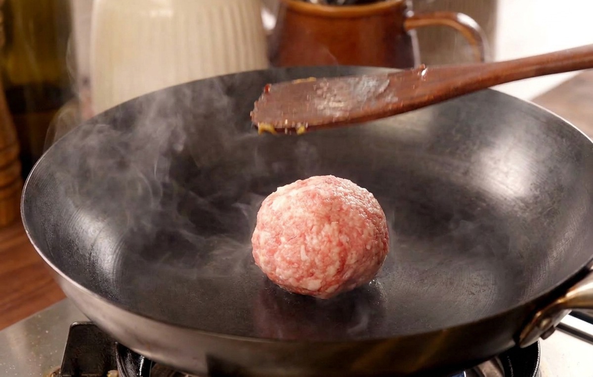 Una spatola di legno che preme con forza su una polpetta di carne in una padella rovente, sprigionando una nuvola di vapore.