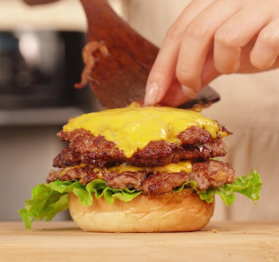 Una persona che usa una spatola per completare un double cheeseburger applicando uno strato di salsa ai peperoni sopra il formaggio fuso.