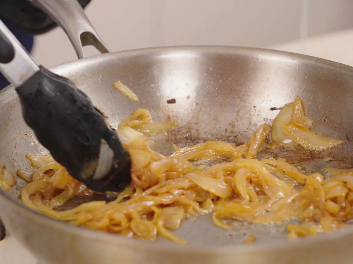 Pinze che mescolano un denso mucchio di cipolle caramellate dorate in una padella di acciaio inossidabile lucida.