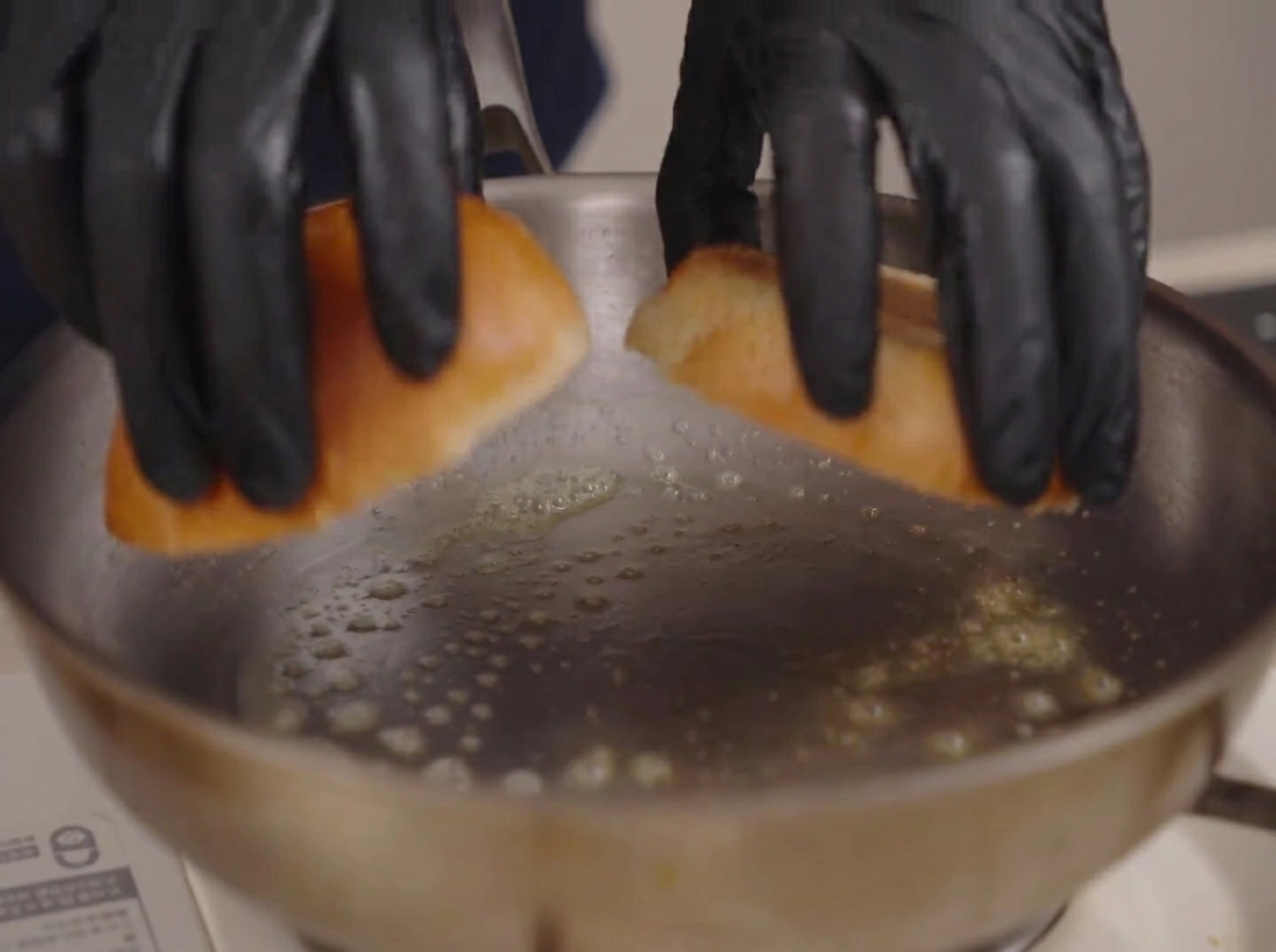 Due mani guantate che posizionano due metà di pane brioche in una padella con burro fuso spumeggiante.