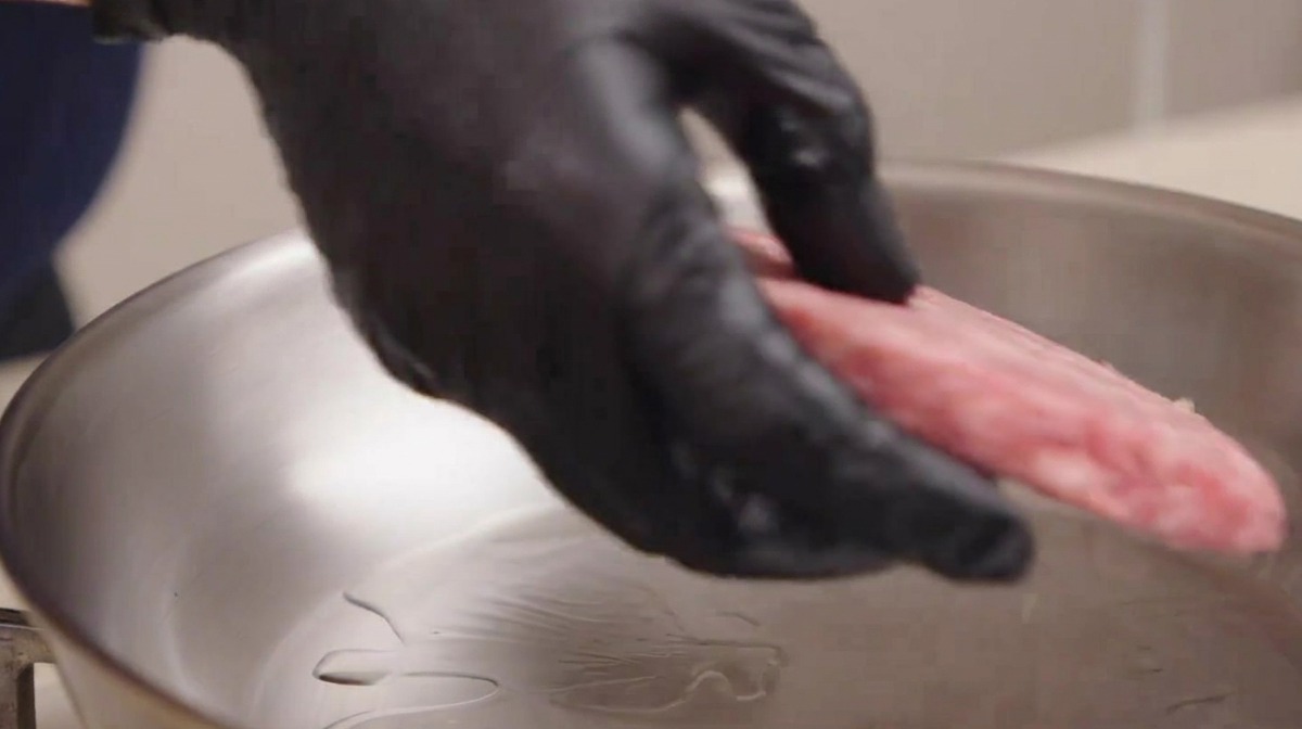 Una mano con guanto nero che posiziona con cura un patty di manzo marezzato crudo in una padella di acciaio inossidabile con olio.