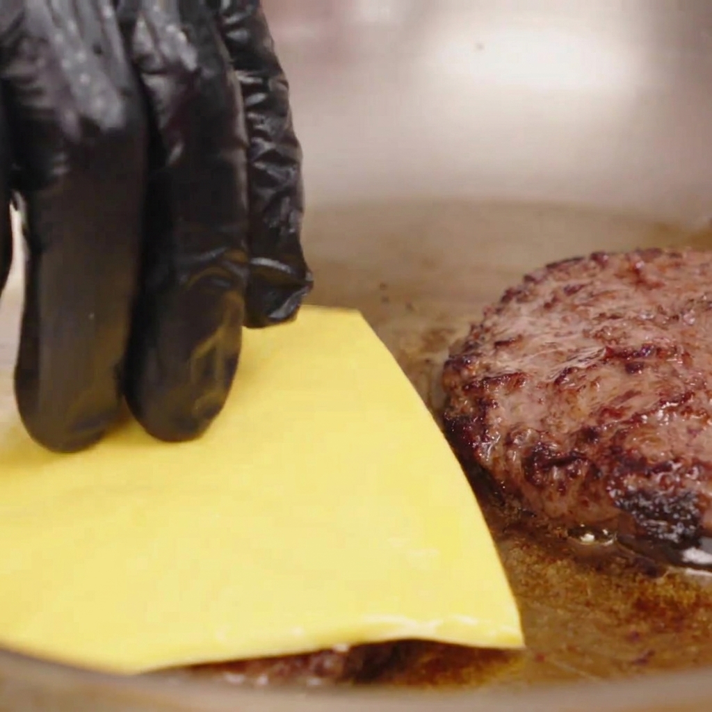 Una mano guantata che posiziona una fetta quadrata di formaggio giallo su un patty di manzo caldo e scottato in padella.
