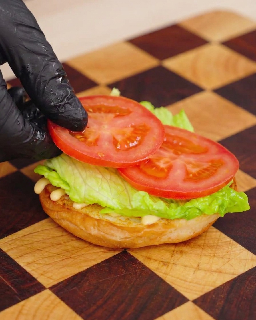 Una mano guantata che posiziona una sottile fetta di pomodoro su uno strato di lattuga verde sopra un panino brioche tostato.