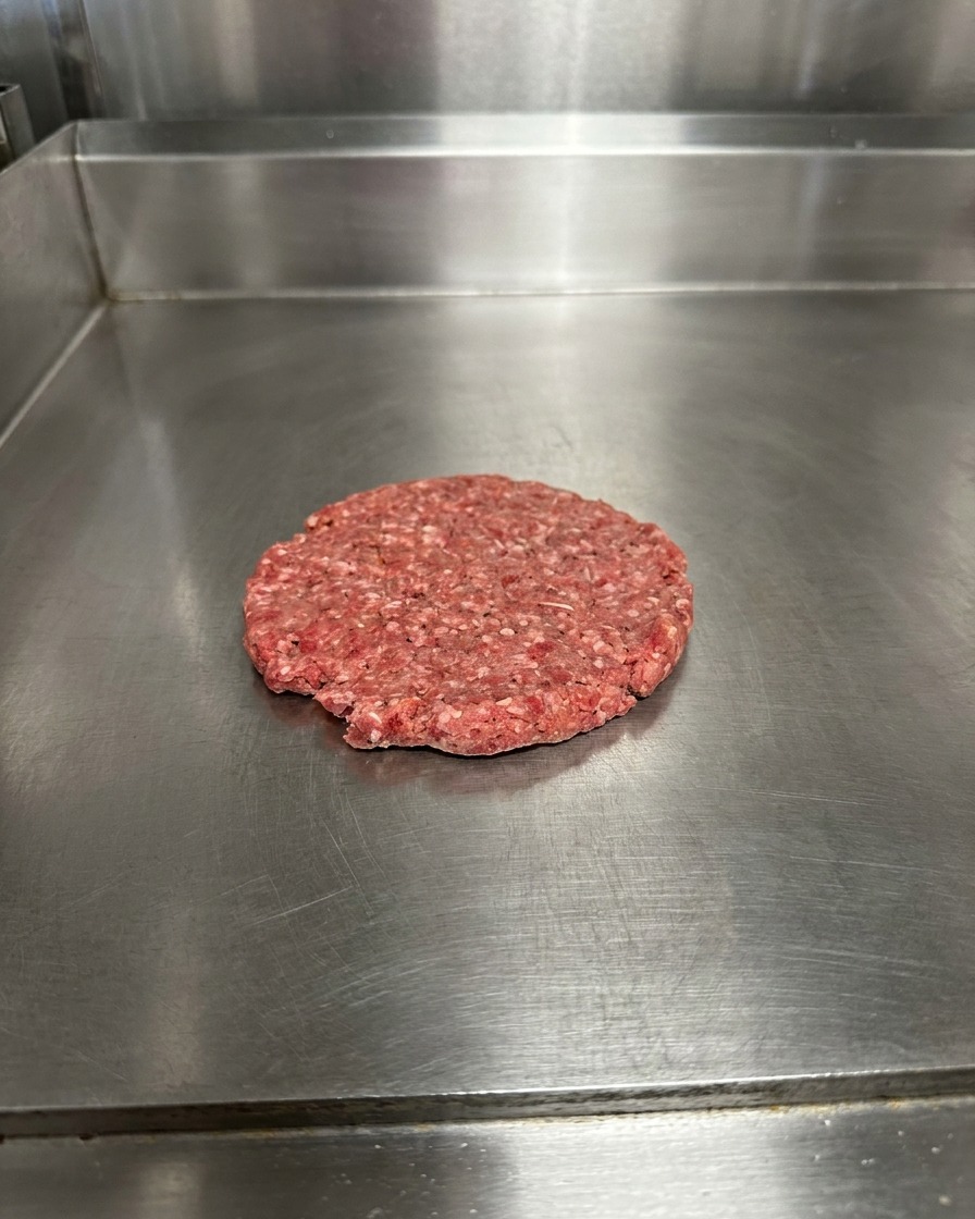 Un patty di manzo schiacciato sulla piastra, condito con grani visibili di sale e pepe.