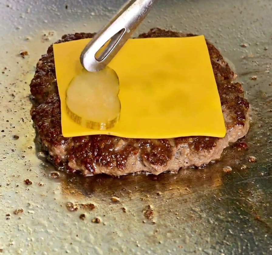 Una fetta di cheddar giallo fuso sul patty mentre vengono aggiunte delle fette di cetriolino con le pinze.