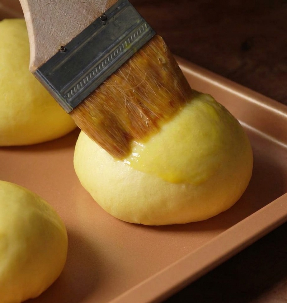 Pennello da cucina che applica luovo sbattuto su un bun lievitato.