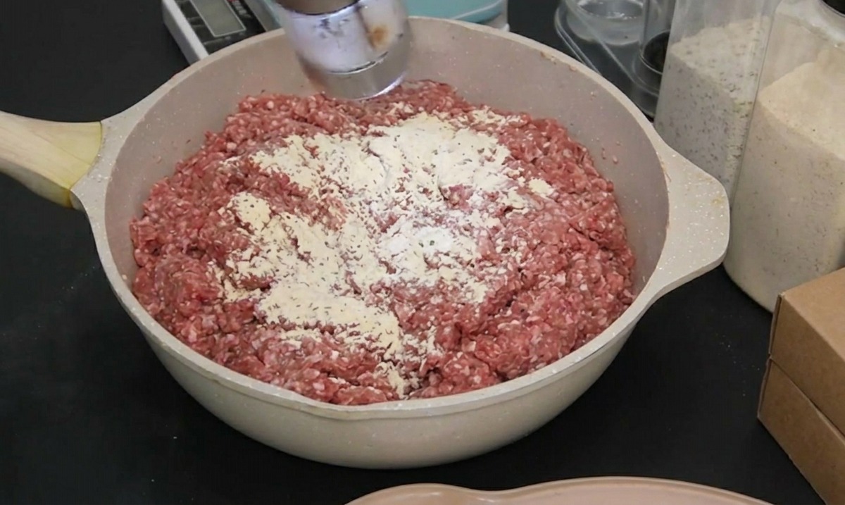 Una persona con un guanto nero usa un macinino elettrico per aggiungere sale marino alla carne macinata cruda.