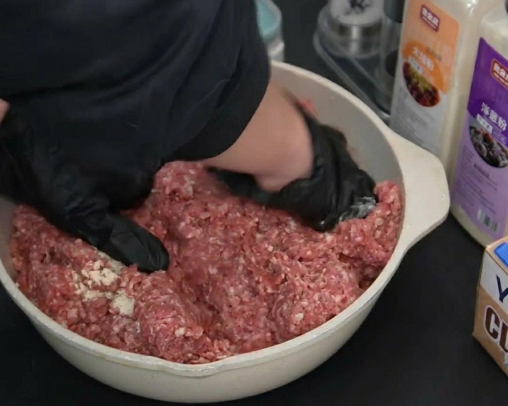 Mani che indossano guanti neri mescolano e impastano la carne macinata cruda condita in una pirofila chiara.