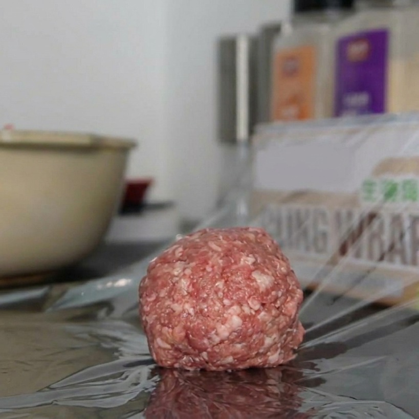 Una singola porzione rotonda di carne macinata condita posata su un pezzo di pellicola trasparente su un bancone.