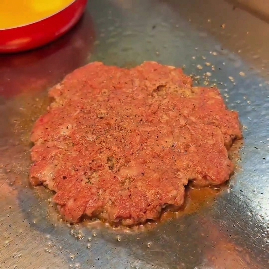Un hamburger di manzo crudo in cottura su una griglia piatta, condito con sale e pepe nero.