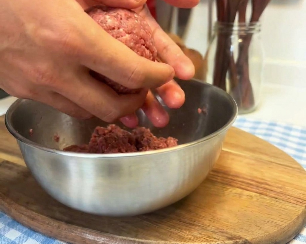 Mani che lavorano una palla di carne macinata cruda sbattendola sul palmo per compattare la consistenza.