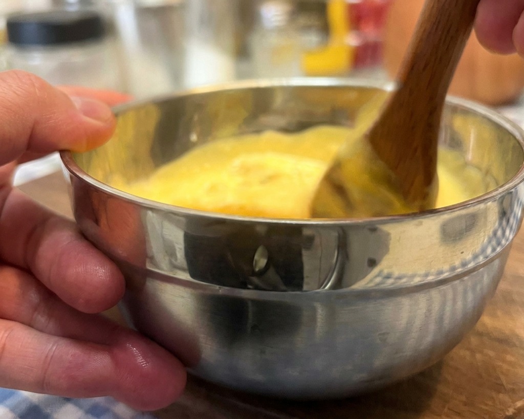 Una mano che mescola una salsa cremosa giallastra con un cucchiaio di legno in una ciotolina di acciaio.
