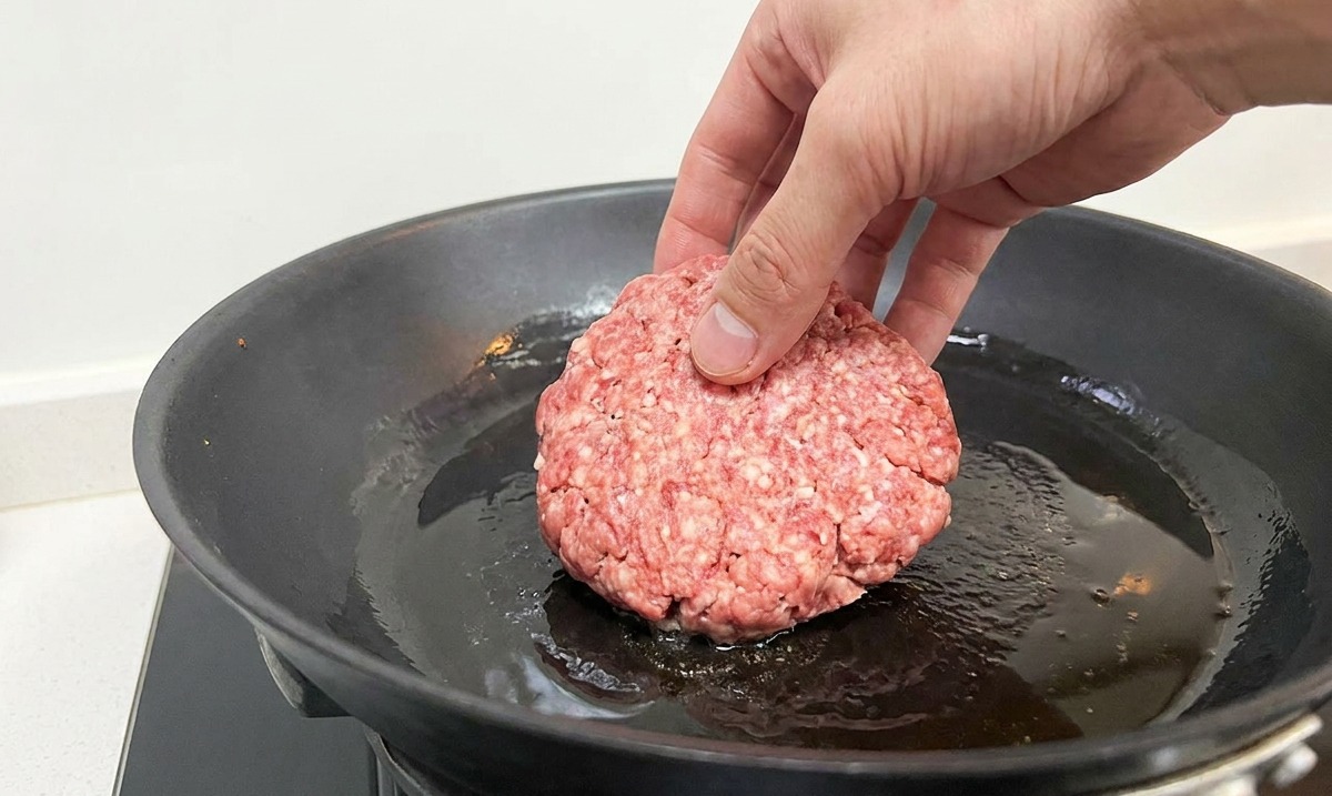 Una mano che adagia un patty di carne cruda in una padella di ghisa calda con olio sfrigolante.
