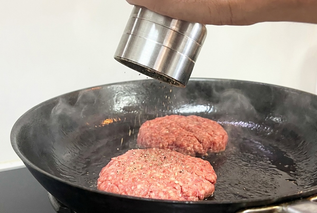 Una mano che usa un macinapepe per condire due patty di carne in padella.