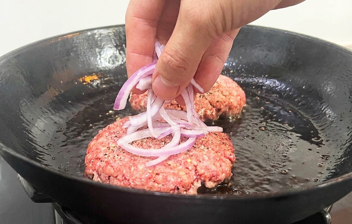Una mano che posiziona fettine sottili di cipolla sopra i patty di carne cruda in padella.