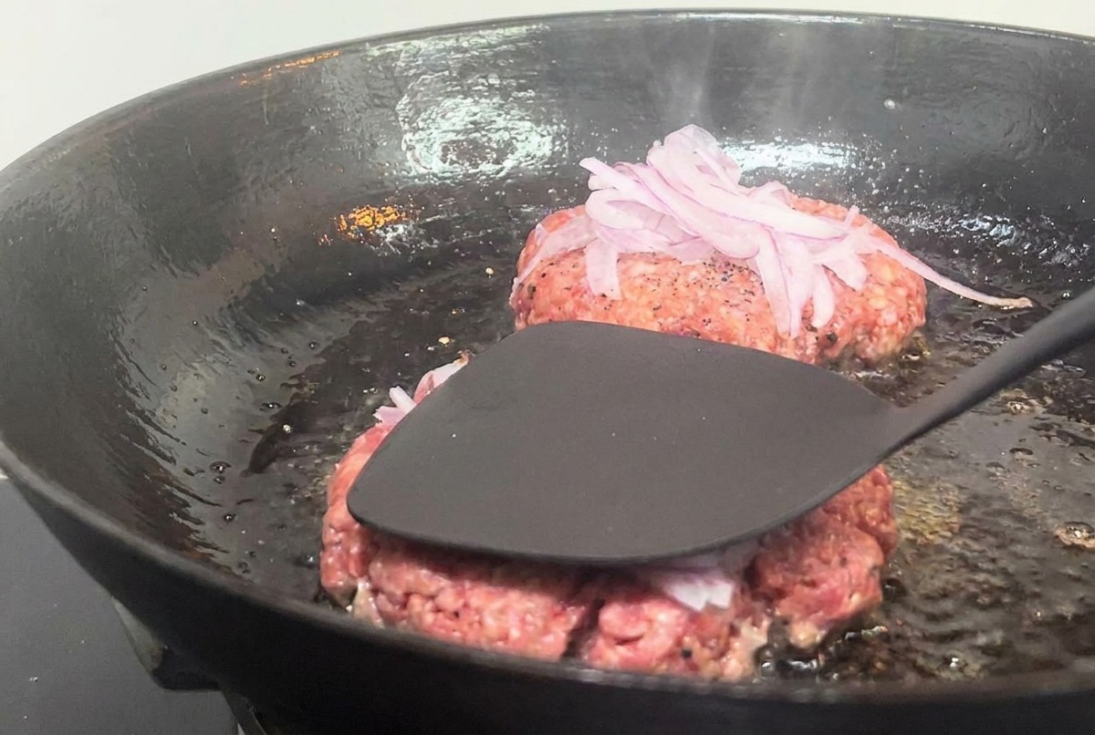 Una spatola metallica che preme con forza su un patty di carne coperto di cipolle in una padella rovente.