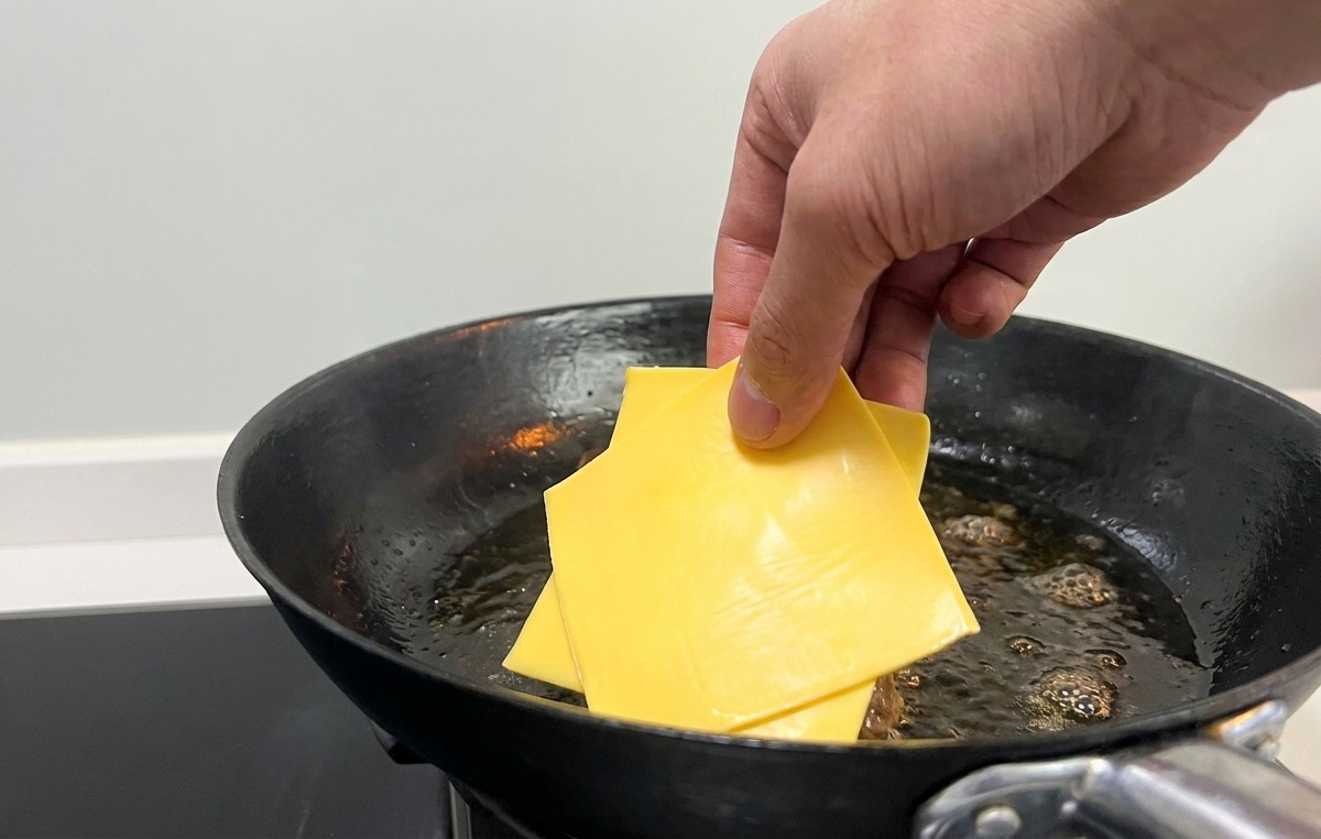 Una mano che appoggia fette di formaggio giallo sopra i patty scottati in padella.