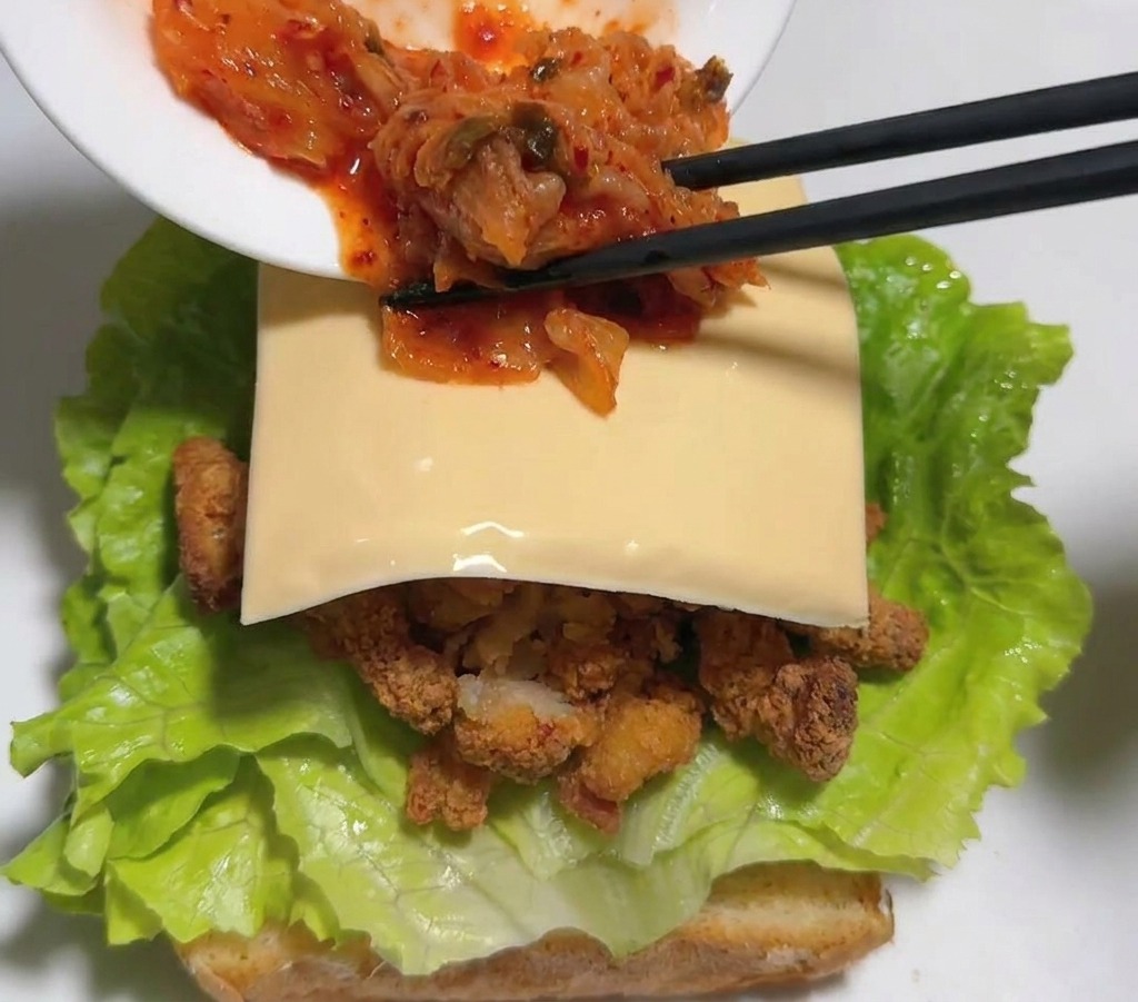 Bacchette nere che trasferiscono kimchi rosso vivace da una ciotola bianca sopra la fetta di formaggio nel sandwich.