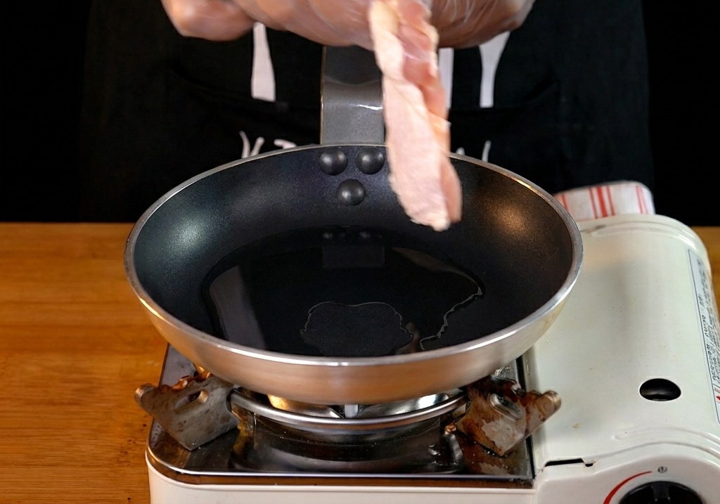 Una mano con guanto trasparente posiziona una sovracoscia di pollo cruda in una padella con olio caldo.