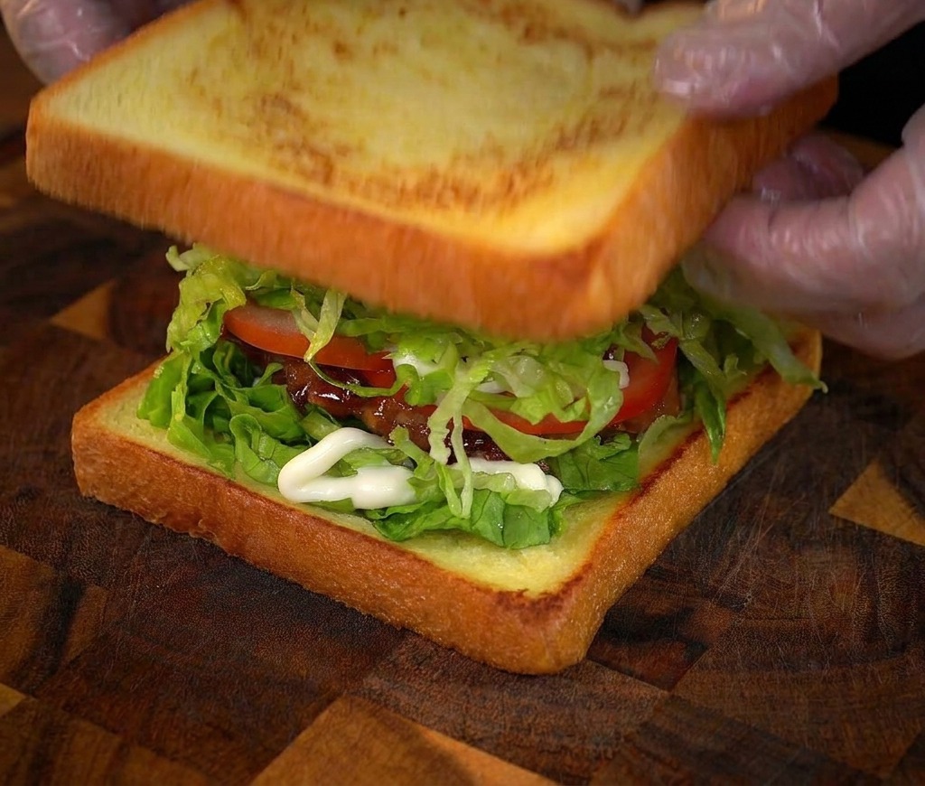 Mani che posizionano lultima fetta di pane tostato sopra un sandwich di pollo alto e stratificato.