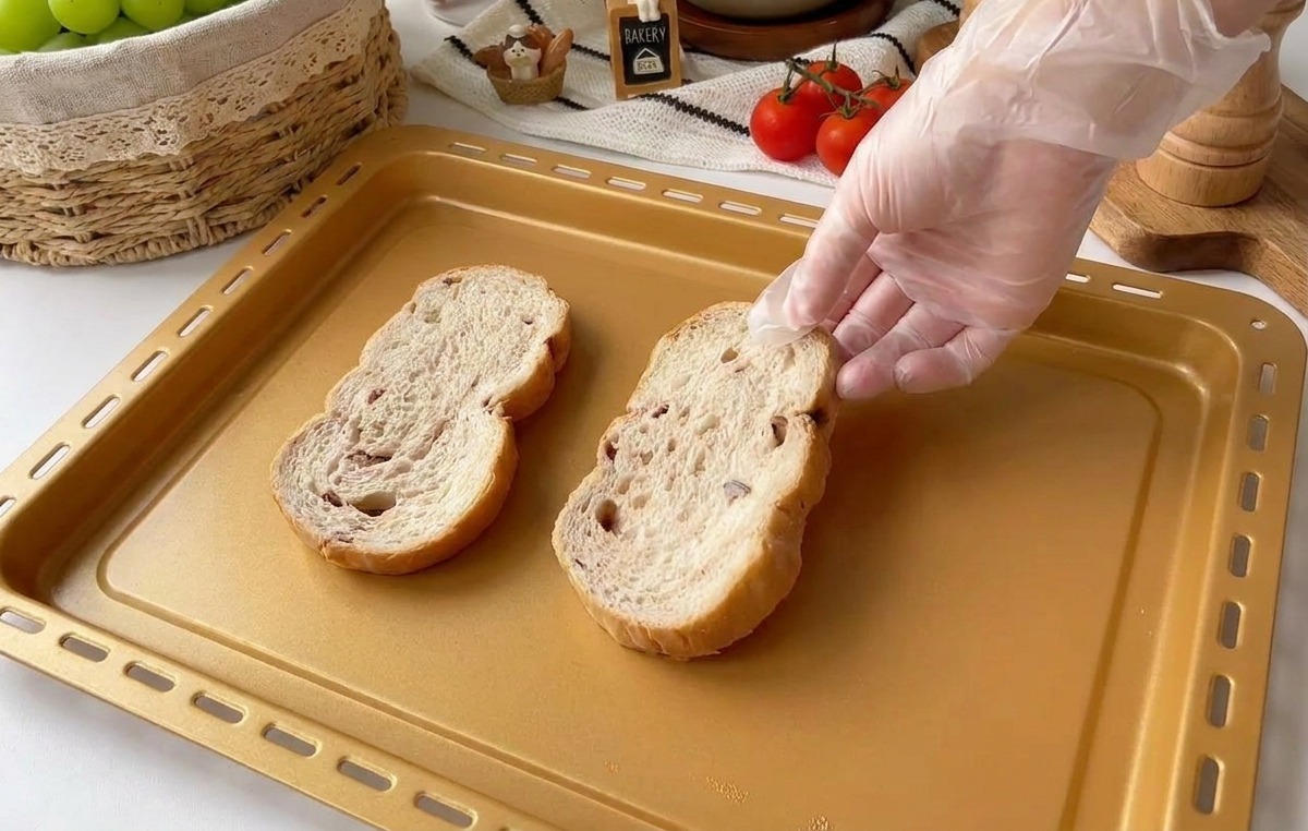 Una mano con un guanto di plastica trasparente posiziona due fette spesse di pane rustico ai cereali su una teglia rettangolare dorata.
