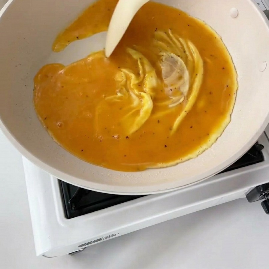 Una spatola bianca muove le uova parzialmente cotte in una padella bianca, creando spesse pieghe gialle e ondulate.