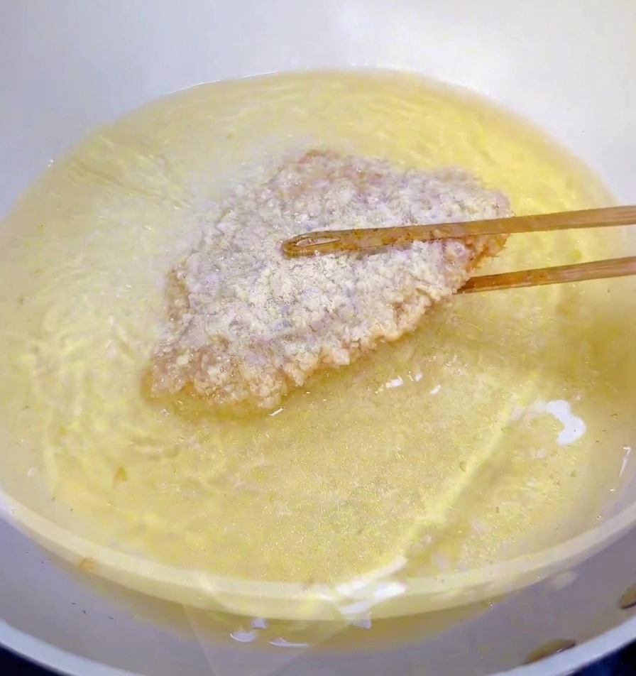 Un filetto di pollo impanato viene immerso in una padella con olio bollente usando bacchette di legno.