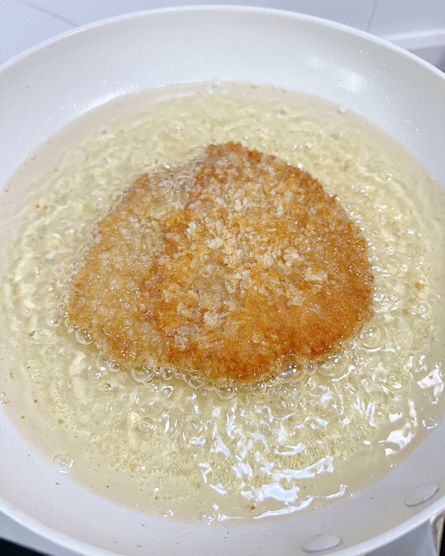 Un filetto di pollo frigge in una padella bianca con olio bollente mentre la crosta diventa dorata e croccante.