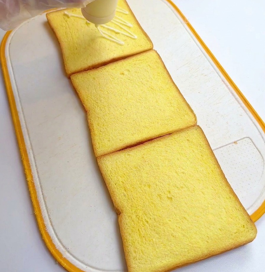 Una persona con guanti di plastica versa del condimento bianco da una bottiglia su tre fette quadrate di pane al latte.