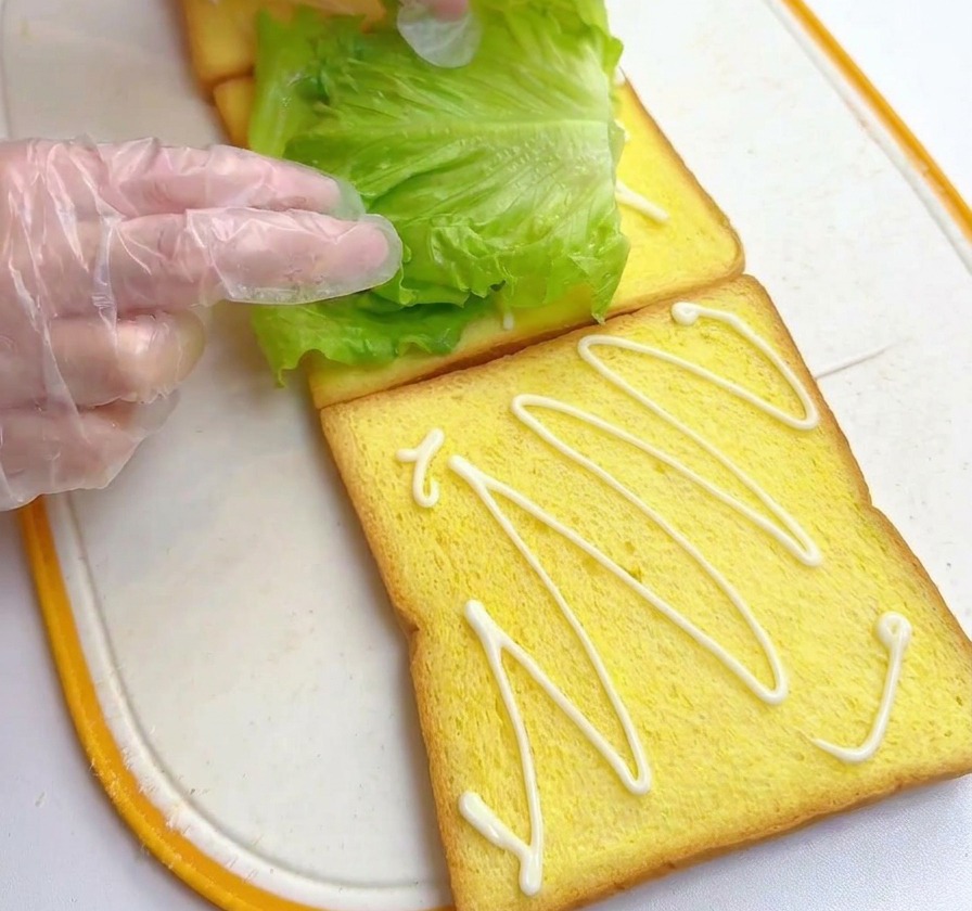 Mani che posizionano una foglia di lattuga verde brillante su una fetta di pane già condita con la salsa.