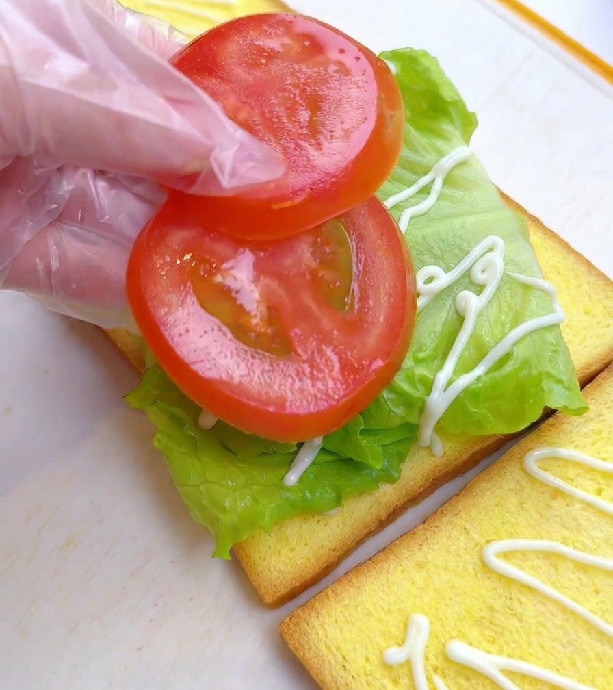 Una persona con un guanto di plastica posiziona due fette tonde di pomodoro su un letto di lattuga verde.