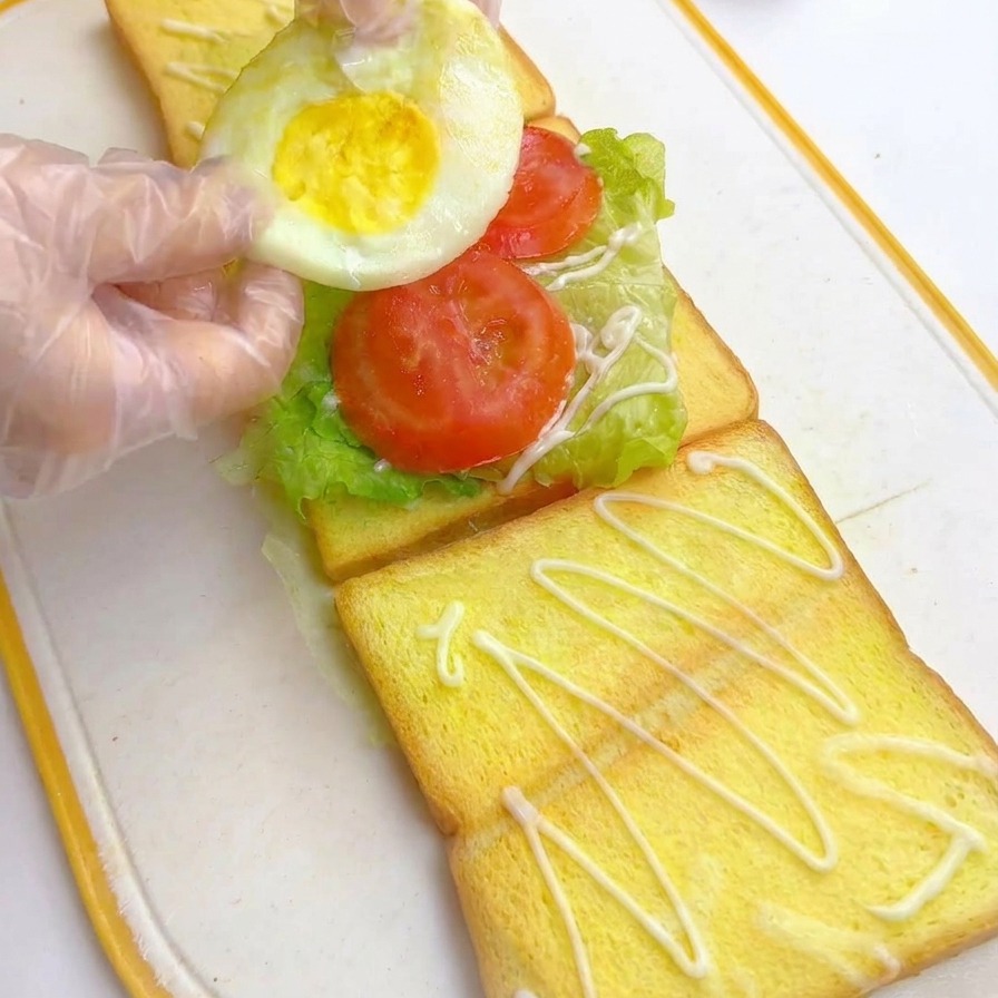 Mani guantate che posizionano un uovo fritto rotondo sopra le fette di pomodoro in un sandwich multistrato.