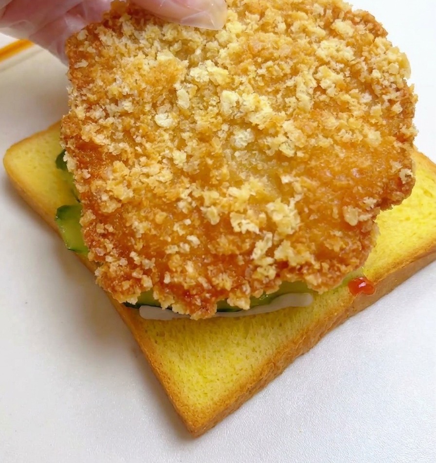 Una mano che posiziona un grande filetto di pollo impanato e fritto sopra la fetta di pane con i cetrioli.