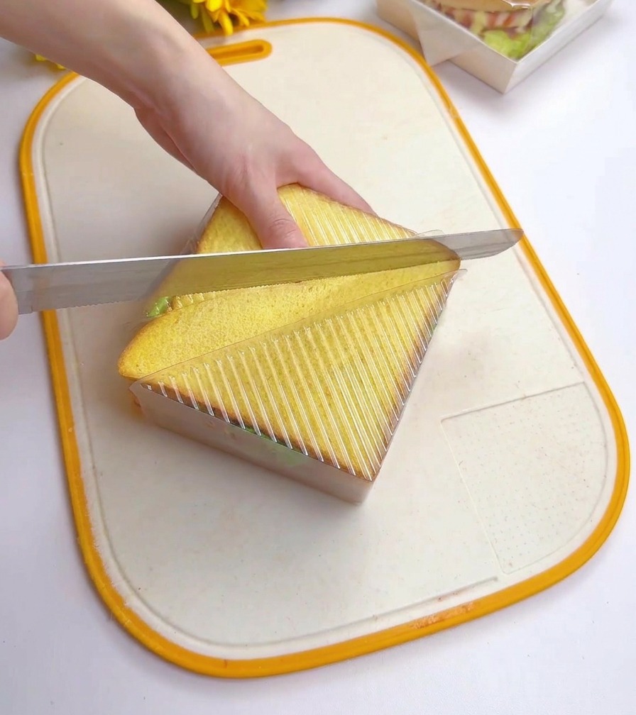 Una persona usa un coltello da pane seghettato per tagliare diagonalmente un sandwich tenuto fermo in uno stampo trasparente.