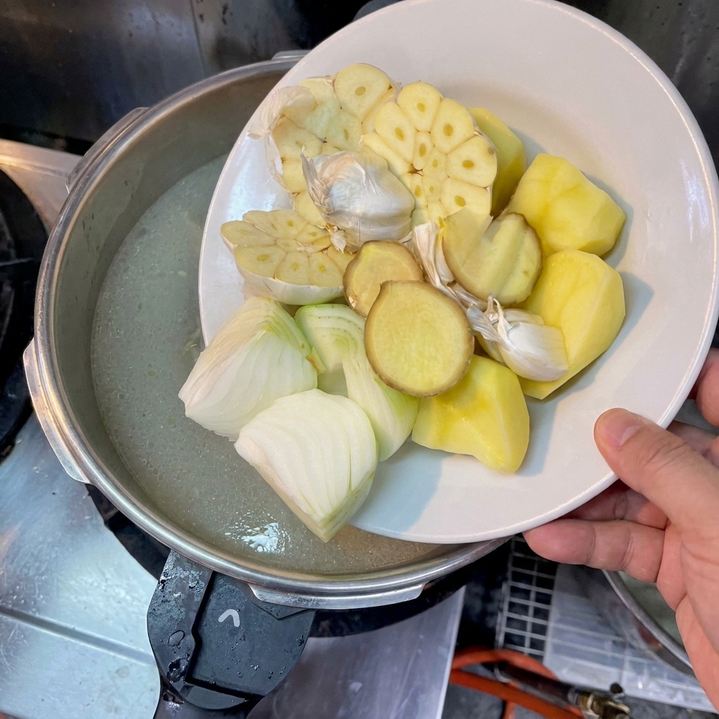 Mano che fa scivolare pezzi di patata, cipolle a metà, zenzero e teste daglio nel brodo.