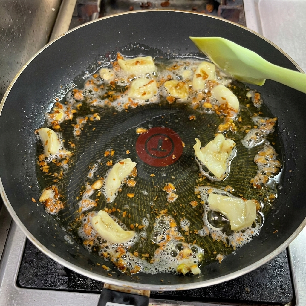 Aglio tritato in padella che diventa dorato scuro mentre viene mescolato con una spatola verde.