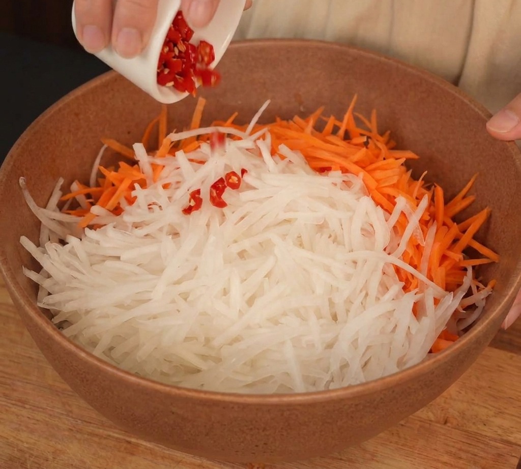 Una ciotola con ravanello bianco e carote a julienne con aggiunta di peperoncini rossi tritati.