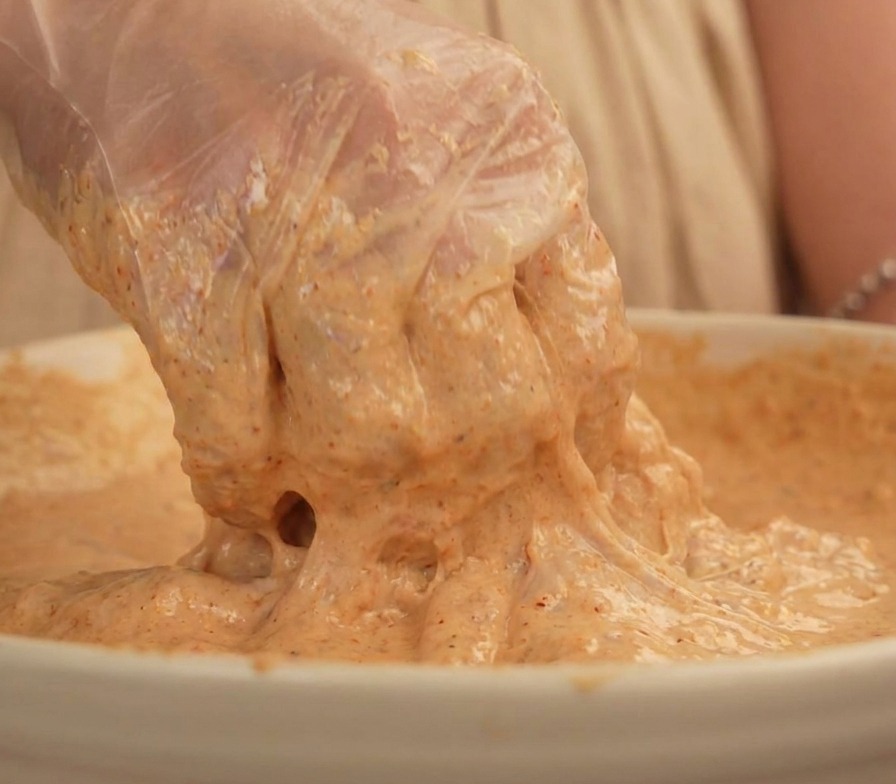 Una mano con guanto solleva un pezzo di pollo completamente avvolto da una pastella densa color beige.