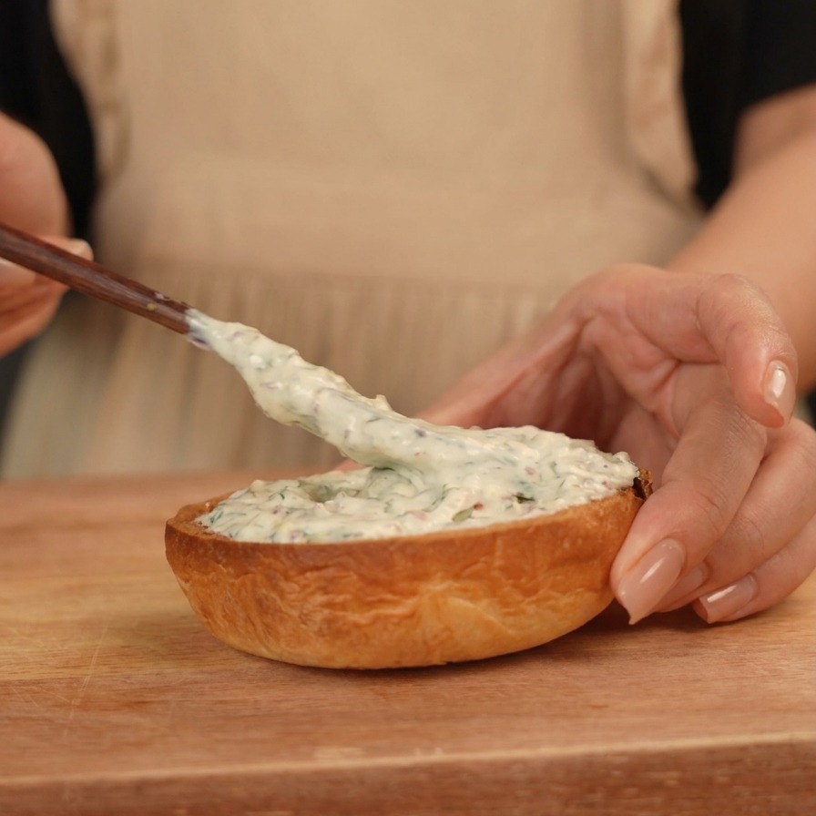 Una mano spalma una salsa bianca cremosa sulla metà inferiore di un panino brioche tostato.
