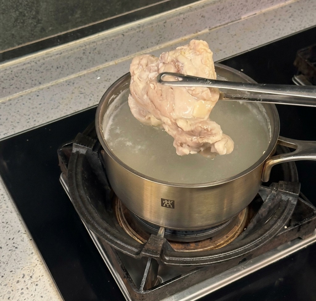 Delle pinze da cucina sollevano un pezzo di pollo cotto da una pentola dacciaio con acqua bollente.
