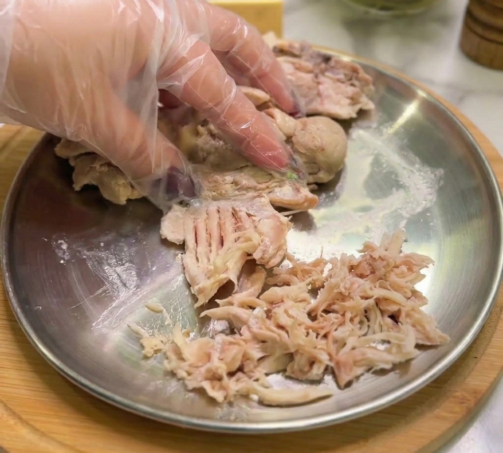 Una mano con un guanto sfilaccia il pollo cotto in strisce sottili su un piatto di metallo.