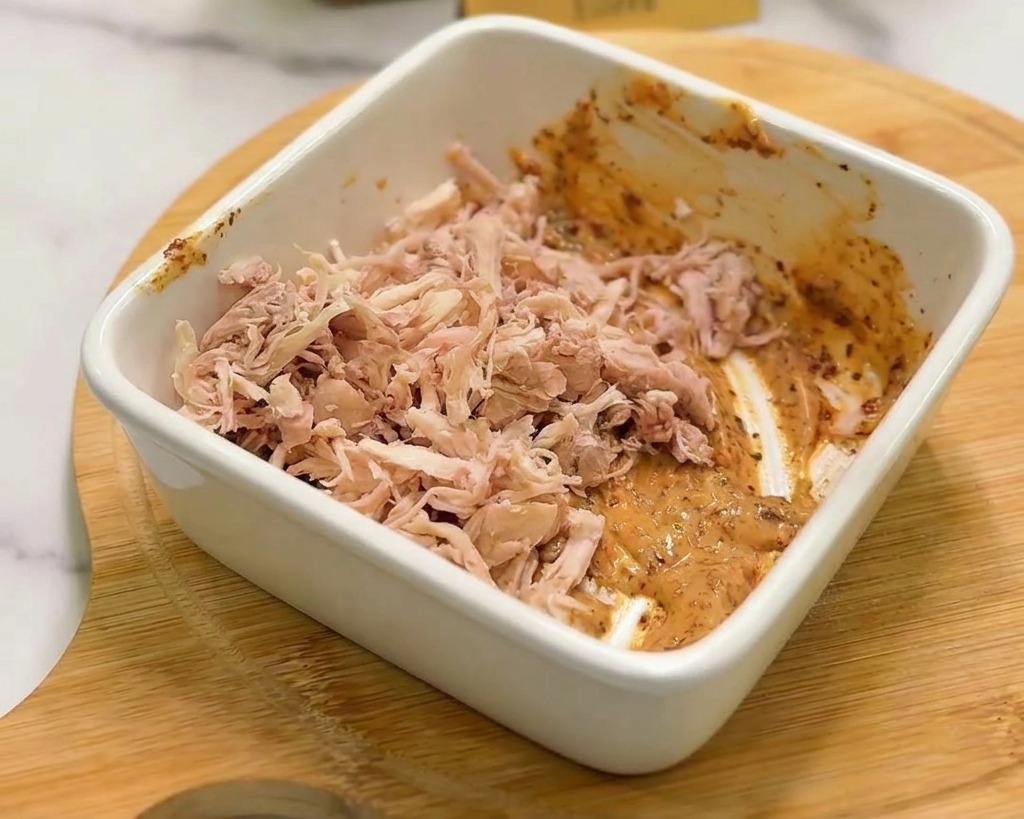 Un mucchietto di pollo sfilacciato aggiunto alla salsa cremosa arancione in una ciotola bianca.