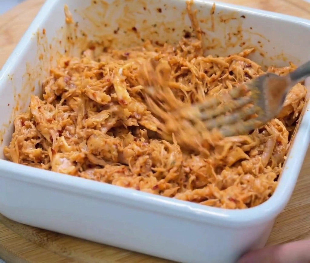 Primo piano di pollo sfilacciato mescolato con una forchetta in una densa salsa rossa allinterno di una ciotola.