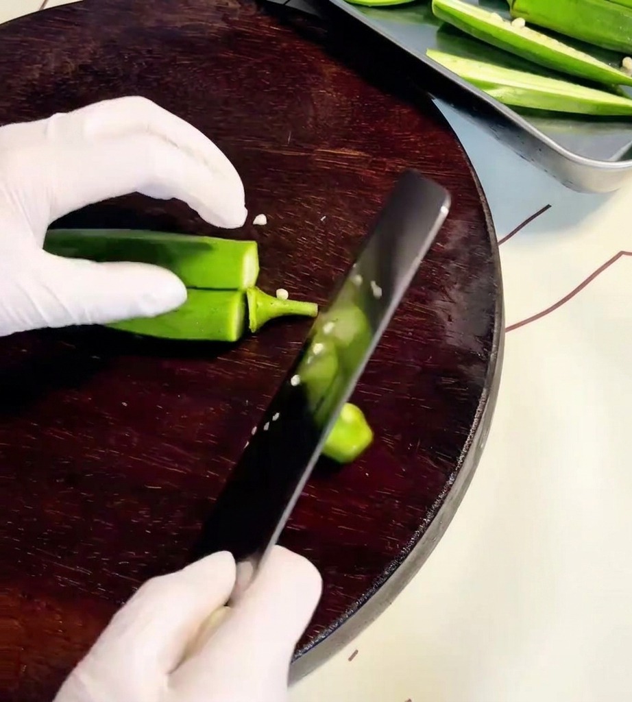Primo piano di mani che usano un coltello per tagliare i baccelli di okra su un tagliere di legno.