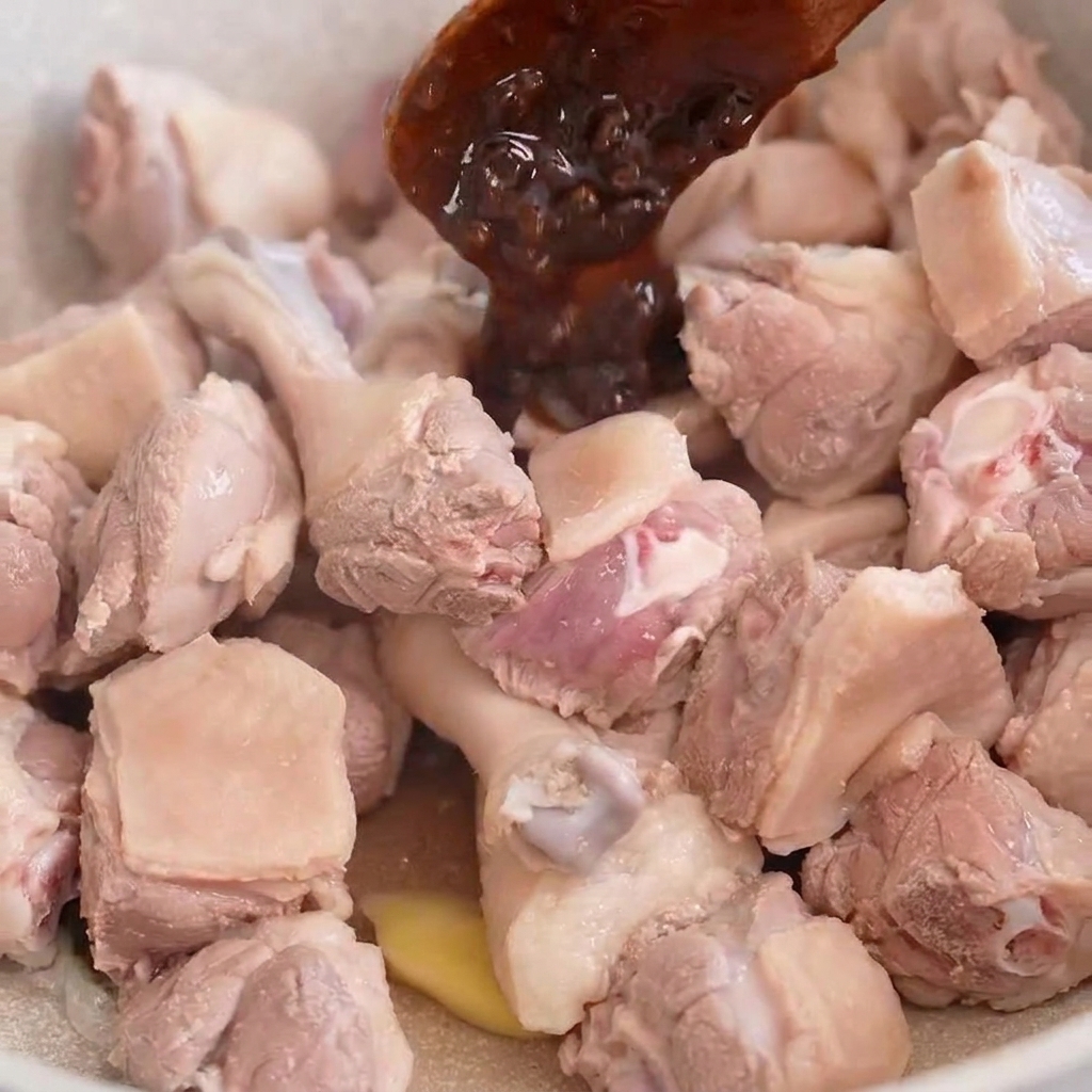 Un cucchiaio di legno che raccoglie pasta di fagioli scura su pezzi di carne danatra che cuociono in una padella dai colori chiari.