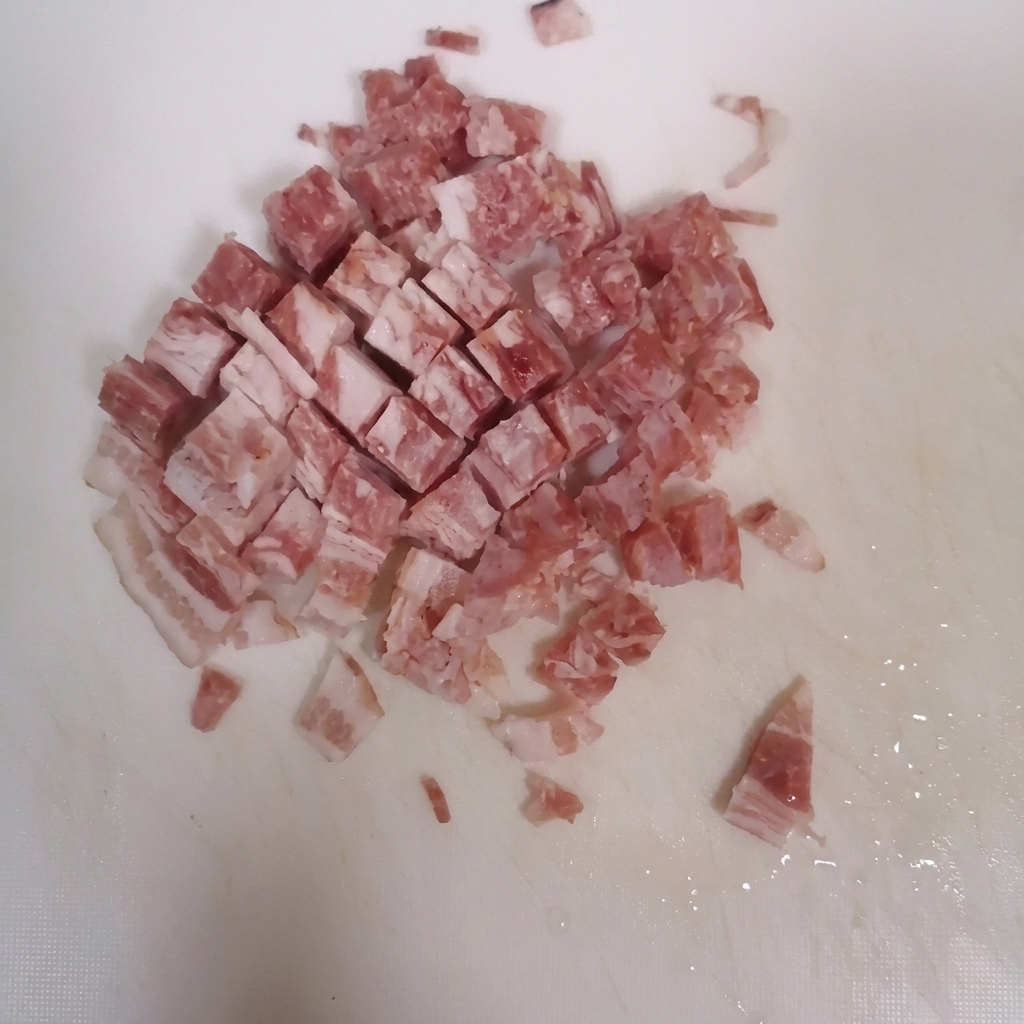 Fette di pancetta tagliate a cubetti su un tagliere bianco pulito.