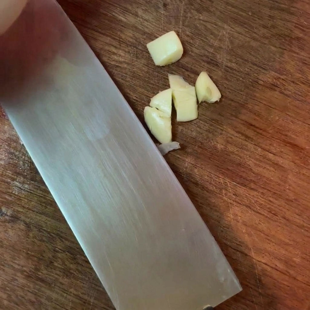 Spicchi daglio tritati grossolanamente accanto a un coltello da cucina su un tagliere di legno.