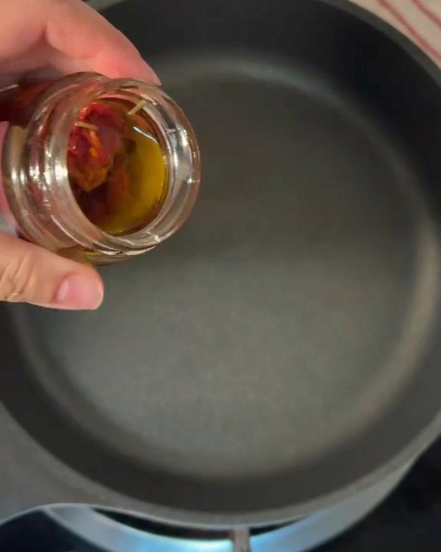 Una mano che inclina un barattolo di vetro per versare olio aromatico ambrato in una padella nera calda.