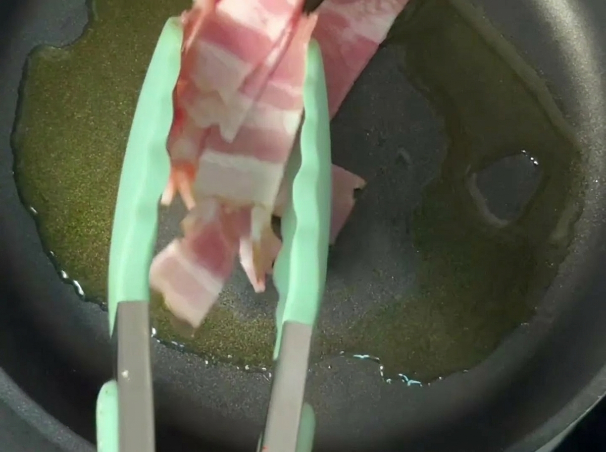 Striscioline di pancetta che sfrigolano nellolio dentro una padella antiaderente.